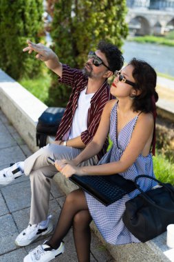 Genç bir çift, güneşli bir günün tadını çıkarıyor. Nehir kenarında oturuyorlar ve bir tablet kullanıyorlar. Şehir ortamında arkadaşlığı ve eğlenceyi gösteriyorlar..