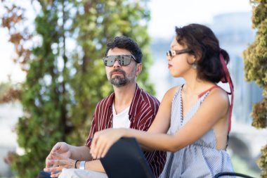 Parkta oturup güneşli bir günün tadını çıkaran şık güneş gözlüğü takan şık bir çift. Bereketli yeşil ağaçlar sakin bir zemin sağlar, rahat ve şık bir ortam yaratır..