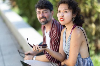 Açık havada sohbet eden iki arkadaş, güneşli havanın tadını çıkarıyorlar. Bir bankta oturuyorlar, yaz kıyafeti giyiyorlar ve güneş gözlüğü tutuyorlar..