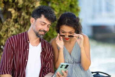 Neşeli bir çift açık havada eğlenceli bir anı paylaşıyor, akıllı telefon ekranına bakıyor, mutluluk ve bağlantı sergiliyor, gündelik, güneşli bir ortamda..