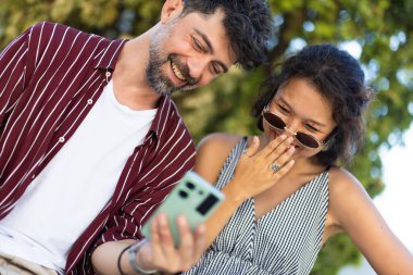 İki arkadaş açık havada akıllı bir telefona bakarken neşeli ve neşeli bir anı paylaşıyorlar, güneşli bir ortamda günlük kıyafetler ve güneş gözlüğü takıyorlar..