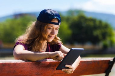 Beyzbol şapkası takan genç bir kadın güneşli bir park ortamında tablet kullanırken gülümsüyor..