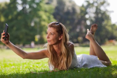 Neşeli genç bir kadın güneşli bir parkta çimenlerde uzanıyor, akıllı telefonuyla selfie çekiyor, bir neşe ve rahatlama anını somutlaştırıyor..