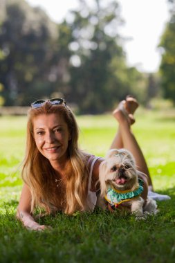 Mutlu Shih Tzu köpeği ile çimenlerin üzerinde uzanmış gülümseyen orta yaşlı bir kadın. Parkta güneşli bir günün tadını çıkarıyor..