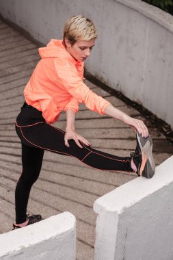 Parlak turuncu spor giysili genç bir kadın ayağını beton basamaklara dayayıp şehir ortamında koşuya hazırlanıyor..
