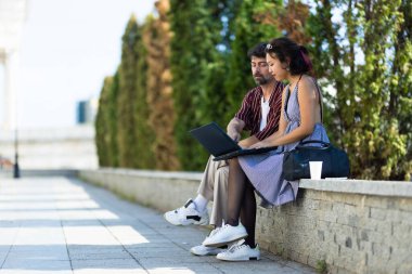 Dışarıda taş bir çıkıntıda oturan genç bir çift laptopta birlikte çalışıyorlar. Odaklanmışlar ve meşguller. Etrafları güneşli bir park ortamında yeşilliklerle çevrili..
