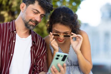 Neşeli bir çift dışarıdaki akıllı telefon ekranına bakar güneşli bir günün tadını çıkarır ve neşeli bir anı paylaşır..