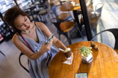 Bir kadın dondurma külahını sever ve rahat bir kafe atmosferinde akıllı telefonuyla zaman geçirir, eğlence ve teknolojinin bir karışımını ön plana çıkarır..