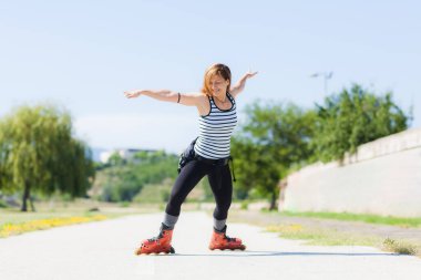 Aktif genç bir kadın parkta paten kayıyor, açık mavi bir gökyüzünün altında çizgili bir askılı bluz ve siyah taytlar giyiyor..
