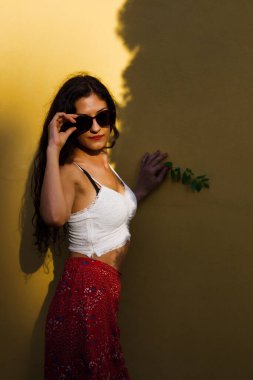 Güneş gözlüğü takan, beyaz bir bluz ve çiçekli bir etek giyen şık genç bir kadın güneşli sarı bir duvarın yanında duruyor..