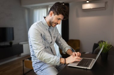 Kot gömlekli odaklanmış genç bir adam modern oturma odasından dizüstü bilgisayarıyla çalışıyor, verimlilik ve konsantrasyonu resmediyor..
