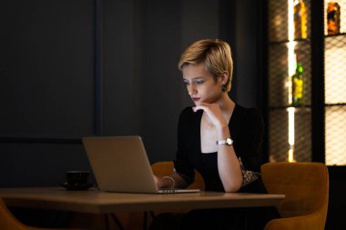 Genç profesyonel bir kadın, modern bir kafe masasında oturmuş, kendini işine adamış ve şık bir çalışma ortamı olan dizüstü bilgisayarına konsantre oluyor..