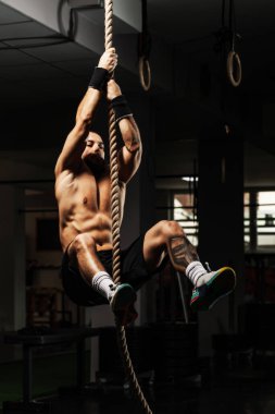 Formda, kaslı bir adam, bir salonda tırmanma ipini kavrıyor. Gücünü ve kararlılığını gösteriyor..