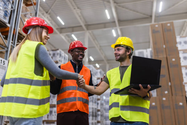 Трое рабочих в спасательных жилетах и шлемах сотрудничают и пожимают руки на складе. Они выглядят вовлеченными и сотрудничающими, окруженными большими стопками коробок и промышленными стеллажами.