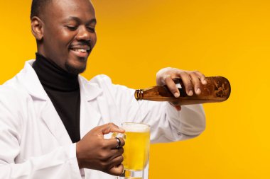 A man wearing a lab coat happily pours beer from a bottle into a glass, set against a vibrant yellow backdrop. clipart