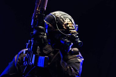 Gece görüş teçhizatı kullanan bir taktik asker karanlık bir ortamda silahla poz vererek, hazırlık ve stratejik askeri operasyonları vurguluyor..