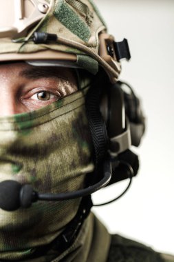 Bir askerin yüzünün yakından görünüşü kısmen taktik teçhizatla kaplı, kamuflaj desenleri, kulaklıklar ve modern askeri teçhizat ve hazırlığın vurgulandığı koruyucu bir miğfer içeriyor..