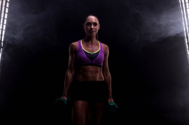Spor kıyafetleri içinde bir kadın, dumanlı arka planı olan loş bir stüdyoda çekilen dambılları kaldırıyor. İfade edici ve ilham verici bir şekilde fitness eğitimine kendini adamış bir güç, güven ve bağlılık sergiliyor..