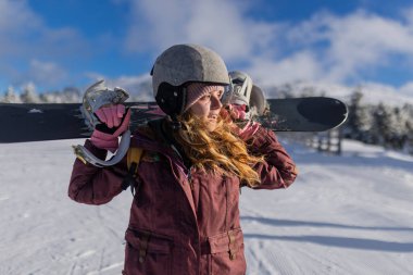 Teçhizat takan ve snowboard 'unu taşıyan bir snowboardcu karla kaplı bir dağ manzarasında duruyor, açık hava kış sporlarının ve canlı bir gökyüzünün altındaki karlı manzaranın tadını çıkarıyor..