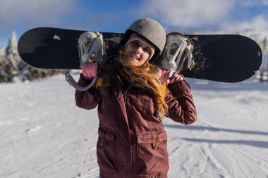 Kış giysileri içinde neşeli bir kadın, omuzlarında bir snowboard taşırken, kış sporları ve açık hava maceraları için heyecanını gösteren karlı bir dağ zeminine doğru gülümsüyor..