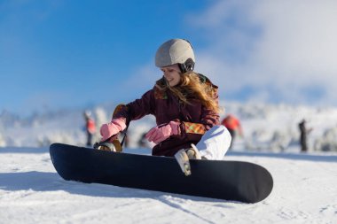 Snowboardcu kış sporlarından zevk alıyor, güzel bir dağ zeminine ve açık mavi gökyüzüne sahip el değmemiş bir kar yamacında oturuyor ve onu hazırlıyor..