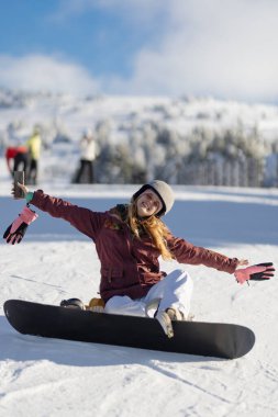 Güzel, karlı bir kış gününde kar kaykayıyla poz veren neşeli genç bir kadın, sakin ve manzaralı bir manzarayla çevrili bir açık hava aktivitesi sergiliyor..