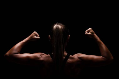 Arkadan kaslı bir kadın kollarını esnetiyor, minimalist karanlık bir arkaplanda güç, sağlık ve beden disiplini temalarını tasvir ederek gücünü ve uygunluğunu vurguluyor..