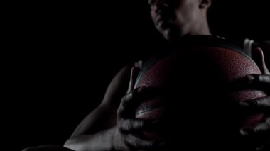 Basketbolcu bankta oturup derin bir nefes alıyor ve aşağıya bakıyor. Siyah arka plana karşı konsantre olmuş Afro-Amerikan sporcu