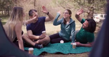 Bir grup genç arkadaş, ormanda çadırın yanında arkadaşlığı ve macerayı sembolize ederken keyifli bir beşlik çakıyor..