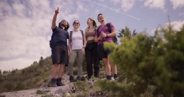 Doğada yürüyüş yapan bir grup arkadaş, uzaktaki ilginç bir şeyi işaret ediyorlar. Gülüyorlar, güneşli günün ve dağ manzarasının tadını çıkarıyorlar..