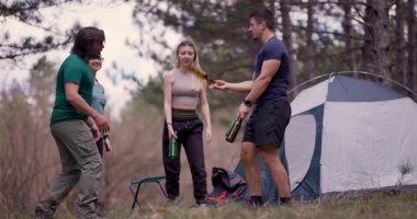 Açık havada giyinmiş genç yetişkinler, bir çadırın yanında bira şişeleriyle hokkabazlık yaparak, doğa gezisinde dostluk ve neşe gösterisi yaparak kutluyorlar..