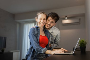 Genç bir adam, kız arkadaşına arkadan sarılır. İkisi de güler ve parlak bir oturma odasında dizüstü bilgisayar kullanır. Konforlu evlerinde neşe ve rahatlığı ifade ederler..