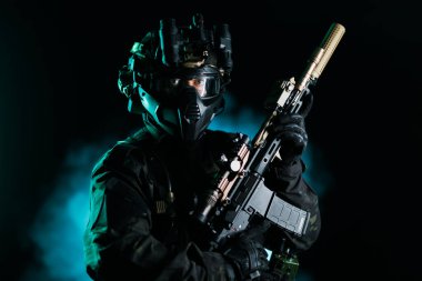 Gelişmiş ekipman giyen, elinde modern bir tüfek tutan ve karanlık arka planda aydınlatma efektleri olan bir taktik şahıs. Modern askeri ve gizli ajanları temsil ediyor.
