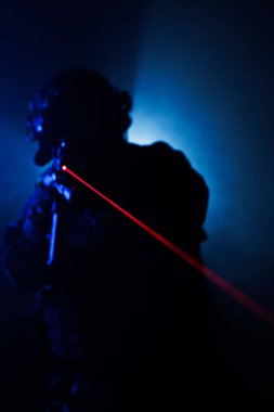 Karanlık, atmosferik bir ortamda, kırmızı lazer ışınlı bir silahı tutan taktik teçhizattaki gölgeli bir figür, gerilim ve yoğunluğu çağrıştırıyor..