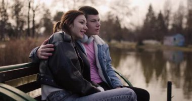 Genç bir çift gölün kenarındaki bankta oturuyor, kulaklıklarını paylaşıyor ve birlikte müzik dinliyorlar..