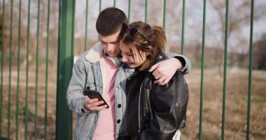 Genç bir çift çitin yanında durup birlikte telefona bakıyor. Sevgiyle sarılırlar, güneşli bir günde birbirlerine eşlik ederek eğlenirler..