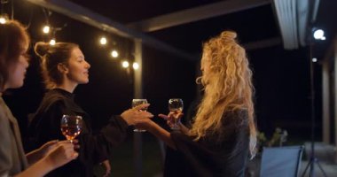 Üç kadın arkadaş, gece terasta içki içip, sicim ışıklarının altında sohbet edip, gülüşüyorlar..