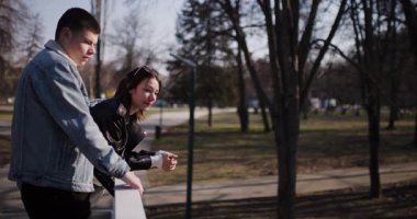 Genç bir çift güneşli bir günde parktaki korkuluklara yaslanır, sohbet eder ve eğlenirler..