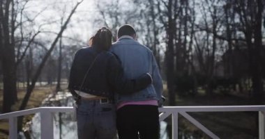 Genç bir çift parktaki beyaz bir köprüde duruyor. Huzurlu manzaranın tadını çıkarıyorlar..