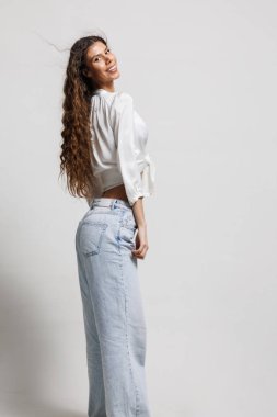 Dalgalı saçlı, parlak gülüşlü sıradan bir kot pantolon ve şık bir bluz giyen mutlu bir genç kadın.