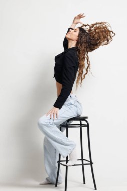 Siyah bluzlu ve açık renk kotlu şık bir kadın dinamik bir poz veren bir taburede oturur, kıvırcık saçlarını ve sanatsal tarzını vurgular..