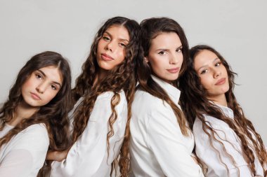 Uzun dalgalı saçlı, beyaz giyinmiş, tarafsız bir arkaplan karşısında stüdyoda yakın poz veren dört genç kadından oluşan bir grup..