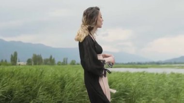 Siyah bir bikini giyen genç bir kadın güneş gözlüğü takıyor ve gölün ve tarlanın yanında dikilirken etrafına bakıyor..