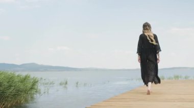 Siyah mayo ve kimono giymiş genç bir kadın, tahta bir iskele boyunca göle doğru yürüyor. Arka planda yavaş çekim.