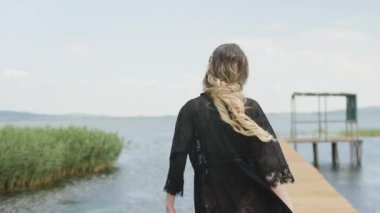 Siyah mayo ve dantelli kimono giymiş genç bir kadın göl kenarında bir terasa doğru yürüyor..