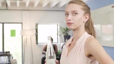 Genç bir kadın modern bir ofiste şişeden su içiyor. Çalışırken su içmek ve dinlenmek için ara veriyor..