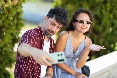 Neşeli genç çift, güneşli bir günde akıllı telefonlarıyla kendi mutluluklarını yakalıyorlar. Günlük yaz kıyafetleri ve güneş gözlüğü takıyorlar, anın tadını çıkarıyorlar..