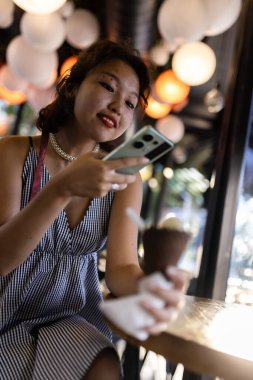 Neşeli genç bir kadın dondurma külahını tutuyor ve canlı, aydınlık bir kafe ortamında akıllı telefonunu kontrol ediyor, günlük eğlenceyi ve modern teknolojiyi somutlaştırıyor..