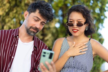 Bir erkek ve bir kadın açık havada güneşli bir günün tadını çıkarıyorlar, akıllı telefona bakıyorlar ve arkadaşça bir anı paylaşıyorlar, arkadaşlık ve teknolojiyi özetliyorlar..