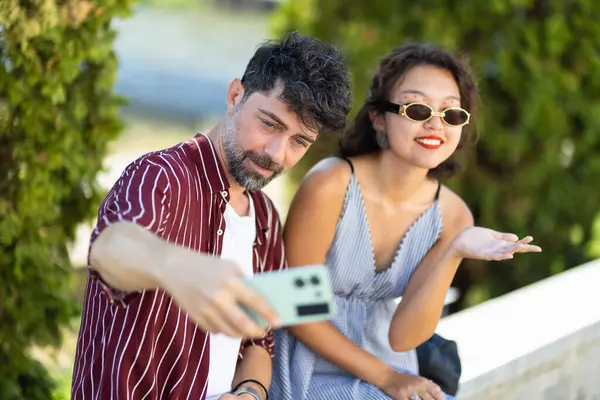 Neşeli genç çift, güneşli bir günde akıllı telefonlarıyla kendi mutluluklarını yakalıyorlar. Günlük yaz kıyafetleri ve güneş gözlüğü takıyorlar, anın tadını çıkarıyorlar..